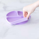 A hand lifts the Lavender Silicone Grip Dish with a secure suction base from Bumkins off a marble surface.