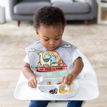 A toddler with a llama bib enjoys fruit and snacks from a Bumkins Silicone Grip Dish in Marble while seated at a high chair.