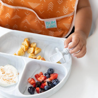 A child in an orange bib enjoys a meal from a Bumkins Silicone Grip Dish: Marble, using a white utensil with pancake, fruit, and yogurt.