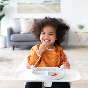 A curly-haired toddler in an orange bib sits happily in a highchair, using a Bumkins Silicone Grip Dish: Marble with suction.