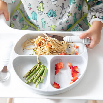 A child enjoys spaghetti, asparagus, and strawberries from a Bumkins Silicone Grip Dish: Marble with a cactus-themed bib and suction base.