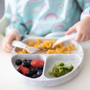 Child in rainbow shirt enjoys pasta, berries, and broccoli from Bumkins Silicone Grip Dish: Marble using fork and spoon on suction base.