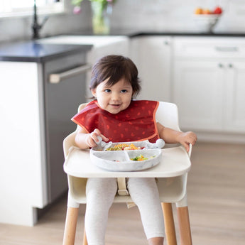 A toddler enjoys food from a Silicone Grip Dish: Marble by Bumkins using a fork, seated in a highchair with a red bib.