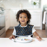 A child with curly hair grins in a bright kitchen, sitting with food in a Marble Silicone Grip Dish by Bumkins and wearing a patterned bib.