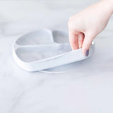 A hand lifts a Bumkins Silicone Grip Dish: Marble, featuring a suction base, from a marble countertop.