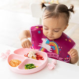 A toddler self-feeds broccoli, strawberries, and applesauce from a Bumkins Silicone Grip Dish: Pink, wearing a unicorn bib.