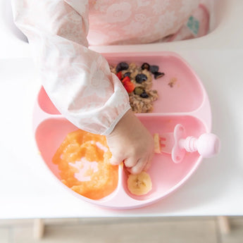 A toddler reaches for food on a Bumkins Silicone Grip Dish: Pink, filled with pureed fruit and oatmeal, at a highchair.