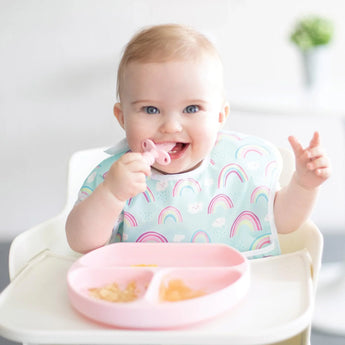 A smiling baby in a high chair wears a rainbow bib and holds a teether with a Bumkins Pink Silicone Grip Dish for self-feeding.