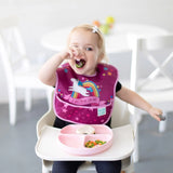 A toddler in a high chair, wearing a unicorn rainbow bib, enjoys self-feeding from a pink Bumkins Silicone Grip Dish on the tray.