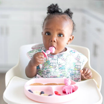 A baby in a high chair enjoys a meal with a pink spoon using the Bumkins Silicone Grip Dish: Pink, featuring divided sections.