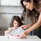 In a bright kitchen, a woman aids a child with their Bumkins Silicone Grip Dish: Pink, filled with berries at the table.