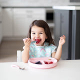 A young girl smiles while sitting at a table with a Bumkins Silicone Grip Dish in Pink, filled with strawberries and blueberries.