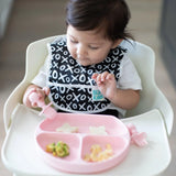 A toddler in a high chair uses a fork with a Bumkins Silicone Grip Dish: Pink, enjoying small food portions.