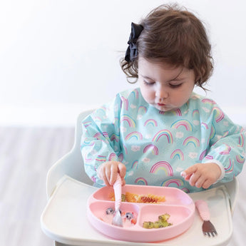 Toddler in a high chair, wearing a rainbow bib, uses a fork to self-feed from a Bumkins Pink Silicone Grip Dish.