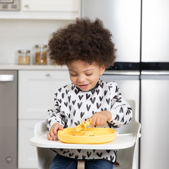 A curly-haired child enjoys toddler portions in a high chair using a Bumkins Silicone Grip Dish: Pineapple, wearing a heart-pattern shirt.