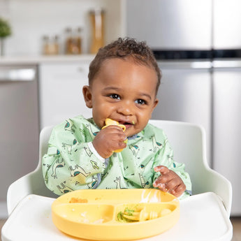 A toddler, in a high chair with a green bib, enjoys their food from a Bumkins Silicone Grip Dish: Pineapple; the kitchen is blurred.