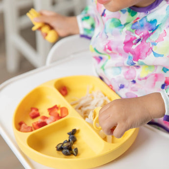 A toddler enjoys pasta, red peppers, & olives from a Bumkins Pineapple Silicone Grip Dish while wearing a colorful shirt. Fun portions!.