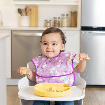 A child in a high chair with a colorful bib enjoys toddler portions from their Bumkins Silicone Grip Dish: Pineapple in the kitchen.