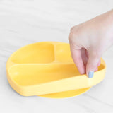 A hand with painted nails holds a yellow Bumkins Silicone Grip Dish: Pineapple, ideal for toddler portions, on a light surface.