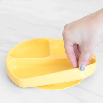 A hand with painted nails holds a yellow Bumkins Silicone Grip Dish: Pineapple, ideal for toddler portions, on a light surface.