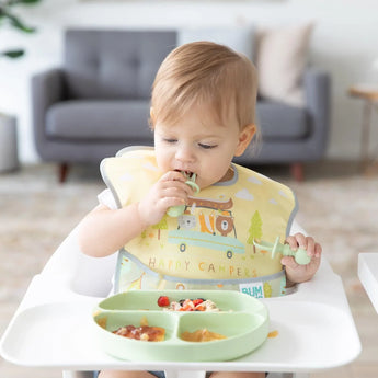 A toddler enjoys a meal in a high chair with a camping-themed bib, holding food from the Bumkins Silicone Grip Dish: Sage.