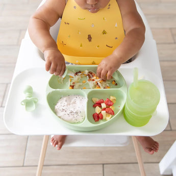 A baby in a high chair enjoys yogurt, chopped fruit, and hash browns on a Sage Silicone Grip Dish by Bumkins with matching cups.