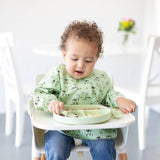 A curly-haired toddler in a green bib plays with food on a Bumkins Silicone Grip Dish: Sage in a high chair, with flowers on the table.