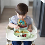 A toddler sits in a high chair, eating pasta and berries from a Bumkins Silicone Grip Dish in Sage. The vibrant bib adds color to the cozy kitchen scene, where the dishs strong suction base keeps it securely in place during mealtime adventures.