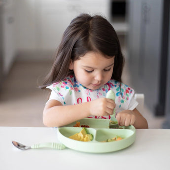 A girl with long hair eats from a Bumkins Silicone Grip Dish in Sage at the table, using a fork. A spoon rests nearby.