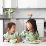 Two kids in matching bibs smile at each other while eating from a Bumkins Silicone Grip Dish: Sage at the kitchen counter.