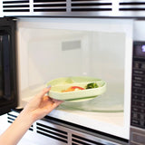 A hand places a Bumkins Silicone Grip Dish in Sage, filled with food, into the microwave—ideal for toddler feeding.