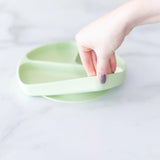 A hand holds the Bumkins Silicone Grip Dish: Sage on a white surface, ideal for toddler feeding.