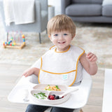 A toddler smiles in a high chair with food on a Bumkins Silicone Grip Dish: Sand, made from food-safe platinum silicone, and a spoon.