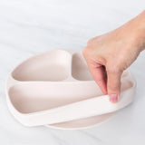 A hand peels the Bumkins Silicone Grip Dish: Sand, a flexible plate with a suction base, made from food-safe platinum silicone.