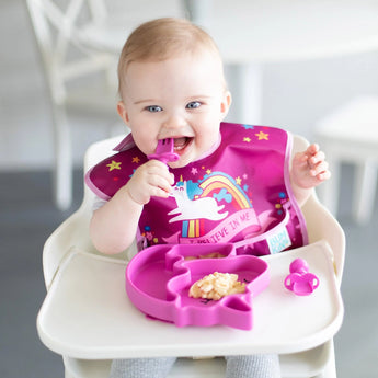 A baby in a high chair enjoys food from a Bumkins Silicone Grip Dish Special Edition: Unicorn, wearing a colorful unicorn bib.