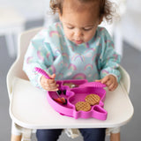 A toddler enjoys waffles and fruit from a pink silicone Unicorn Grip Dish by Bumkins while sitting in a highchair.