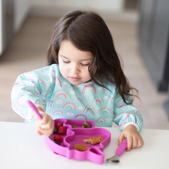 A child happily snacks using the Bumkins Unicorn Silicone Grip Dish, featuring a pink unicorn design and rainbow bib.