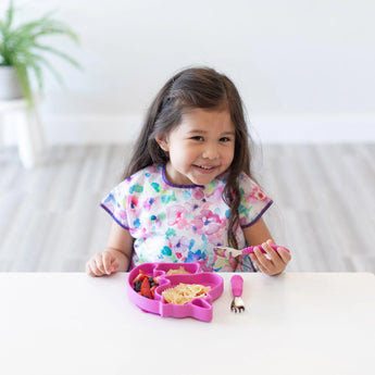 Young girl smiles indoors at table, eating fruit and pasta from Bumkins Silicone Grip Dish Special Edition: Unicorn.