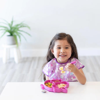 Girl in a purple bib smiles, using a pink fork with her Bumkins Unicorn Silicone Grip Dish. Plant in the background.