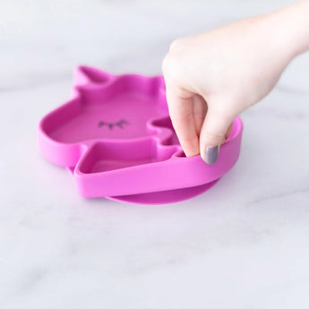 Hand lifting the Bumkins Unicorn Silicone Grip Dish on a white surface.