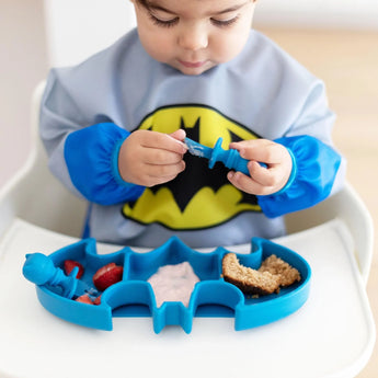 A child in a superhero bib plays with a blue spoon over a Bumkins Batman Blue Silicone Grip Dish filled with strawberries, yogurt, and bread.