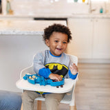 A toddler enjoys snacks in a high chair with the Bumkins Batman Blue Silicone Grip Dish, ideal for little superheroes.