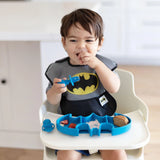 A toddler in a Batman bib enjoys snacks from a blue Bumkins Silicone Grip Dish: Batman Blue while seated in a high chair.