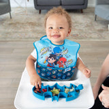 A smiling baby in a superhero bib enjoys food on a Bumkins Batman Blue Silicone Grip Dish.