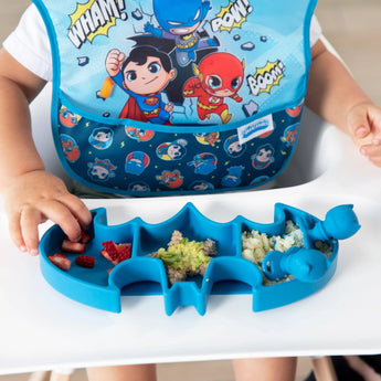 A child enjoys snacks from a Bumkins Silicone Grip Dish: Batman Blue, wearing a superhero-themed bib at the high chair.