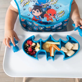 A child enjoys finger foods on a Bumkins Silicone Grip Dish: Batman Blue, wearing a superhero-themed bib at the highchair.