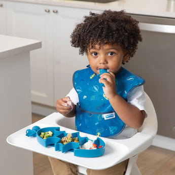 A toddler enjoys snacks from the Bumkins Silicone Grip Dish: Batman Blue, wearing a matching bib, seated in a highchair in the kitchen.