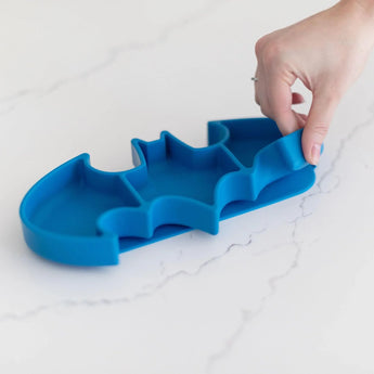 A hand lifts a Bumkins Batman Blue Silicone Grip Dish on a white marble surface.