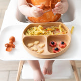 A baby in a highchair, wearing an orange bib, holds a spoon and enjoys pasta, crackers, and fruit from Bumkins Wood Grain Silicone Grip Dish.