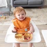 A toddler in a high chair enjoys Bumkins Silicone Grip Dish: Wood Grain with food-safe silicone and suction base, wearing an orange bib.